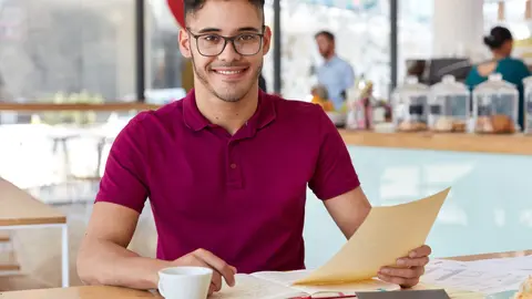 Smiling datisfied entrepreneur holds papers, wears casual clothes, prepares for training workshop, reads necessary information, analyzes documentation, poses against cafe interior. Working conditions